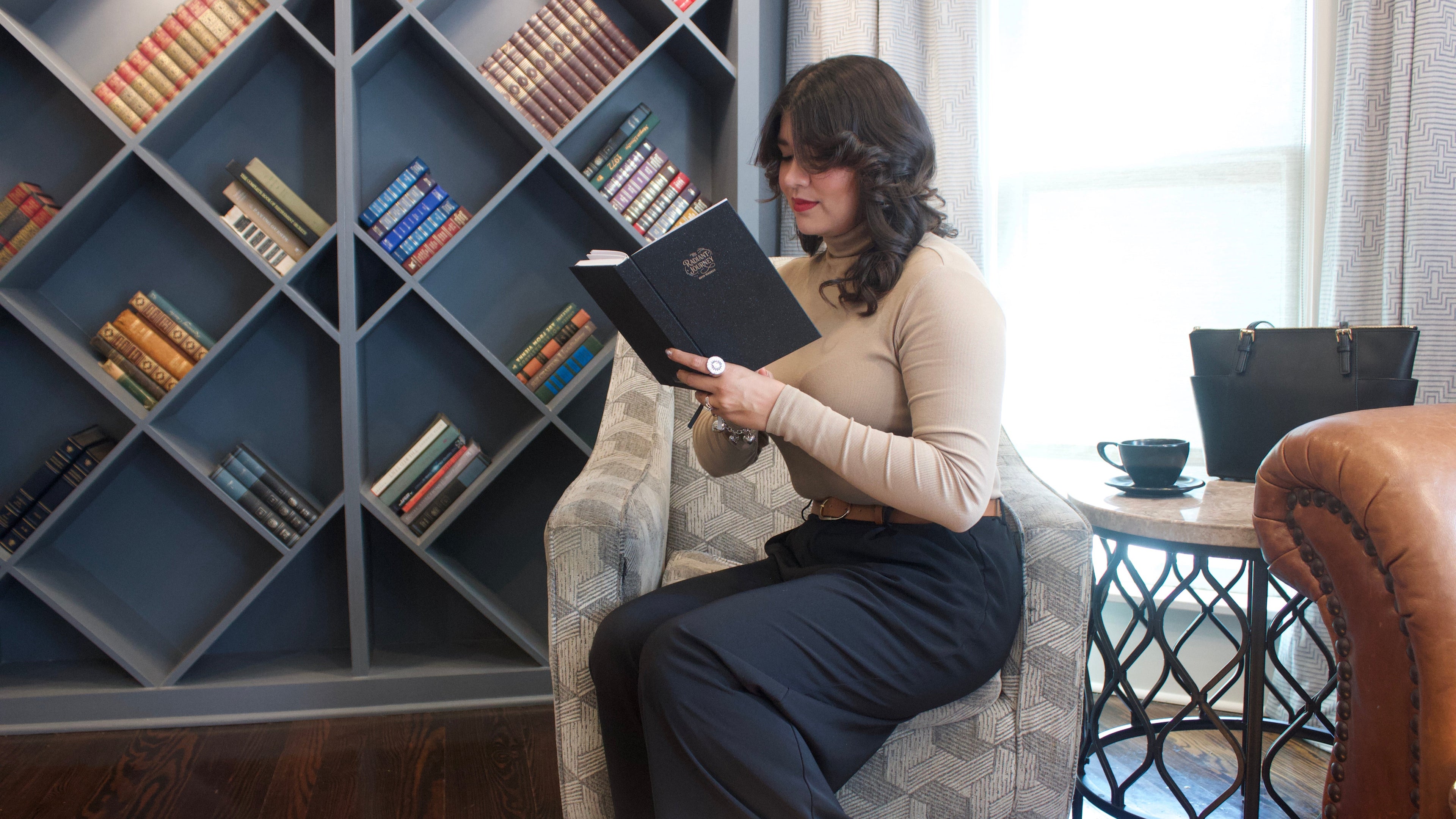 Woman reading a book in a cozy room with a bookshelf and table.
